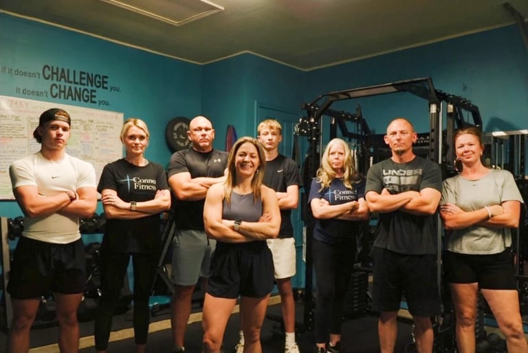 Team of eight fitness enthusiasts posing together in a gym with arms crossed, wearing matching dark athletic wear against a turquoise wall.