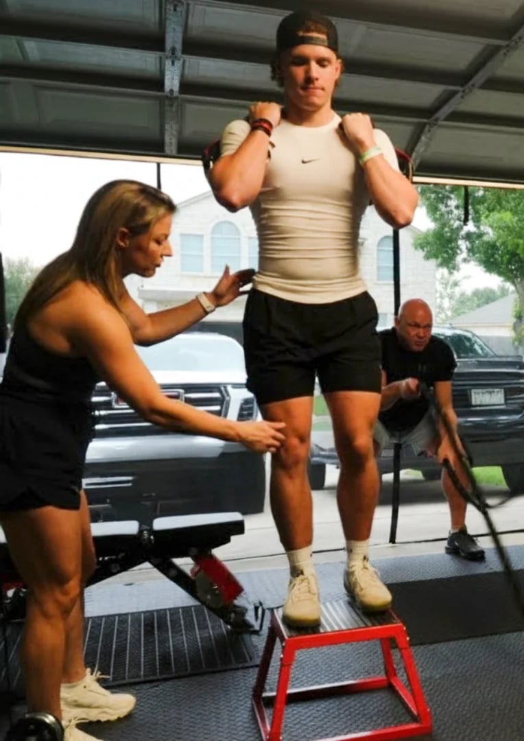 Male athlete performing a box jump exercise on a red plyometric box while female trainer observes in an outdoor gym setting